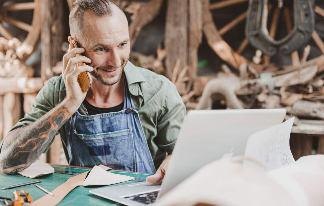 Artisan travaillant sur ordinateur portable et téléphone dans son atelier pour gérer son activité et sa présence en ligne.